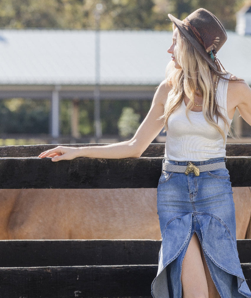 Woman in a white tank top and blue denim skirt standing next to a horse, wearing a brown hat.